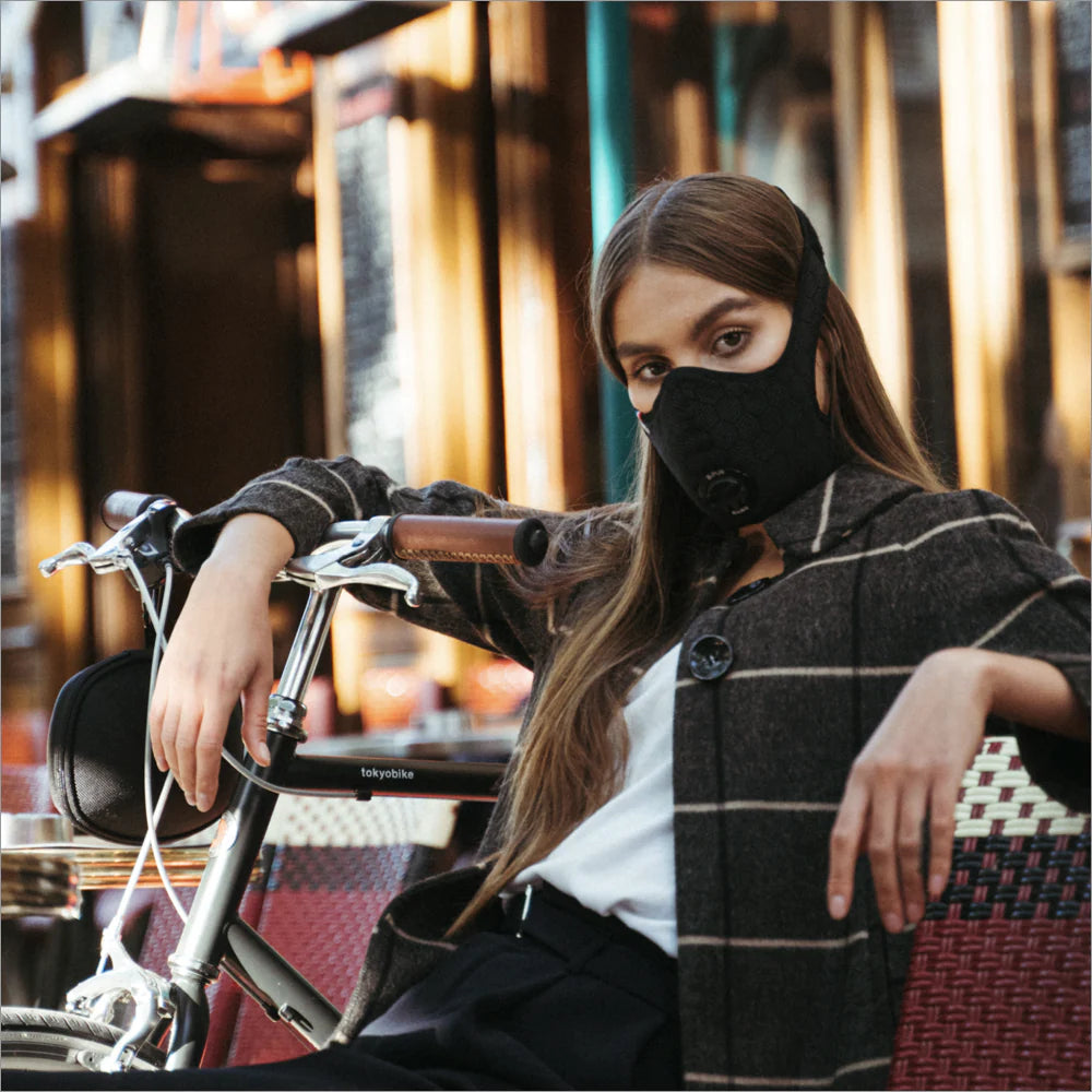 Woman relaxing by a city bicycle at a café while wearing a black R-PUR mask, an urban travel mask look