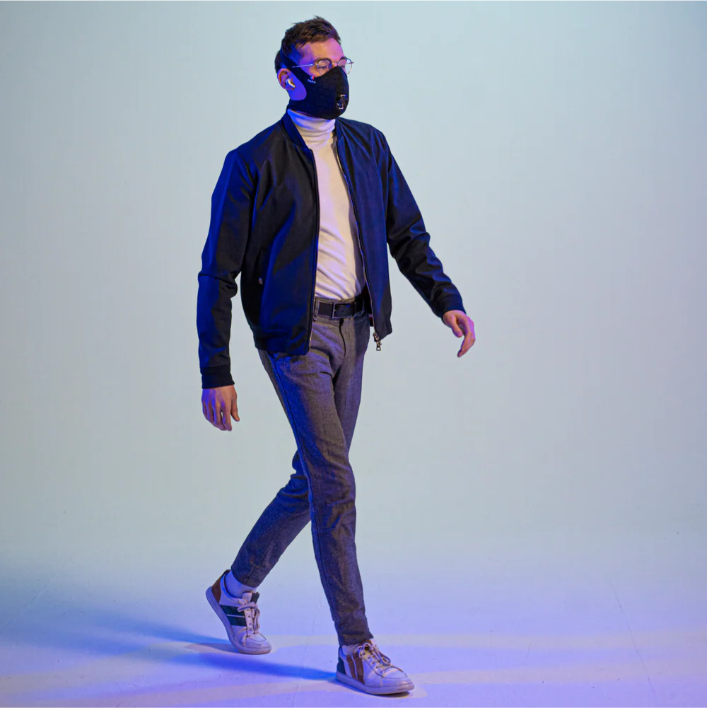 man walking while wearing a travel anti-pollution mask, a cap, and headphones, with his arms crossed and looking to the side