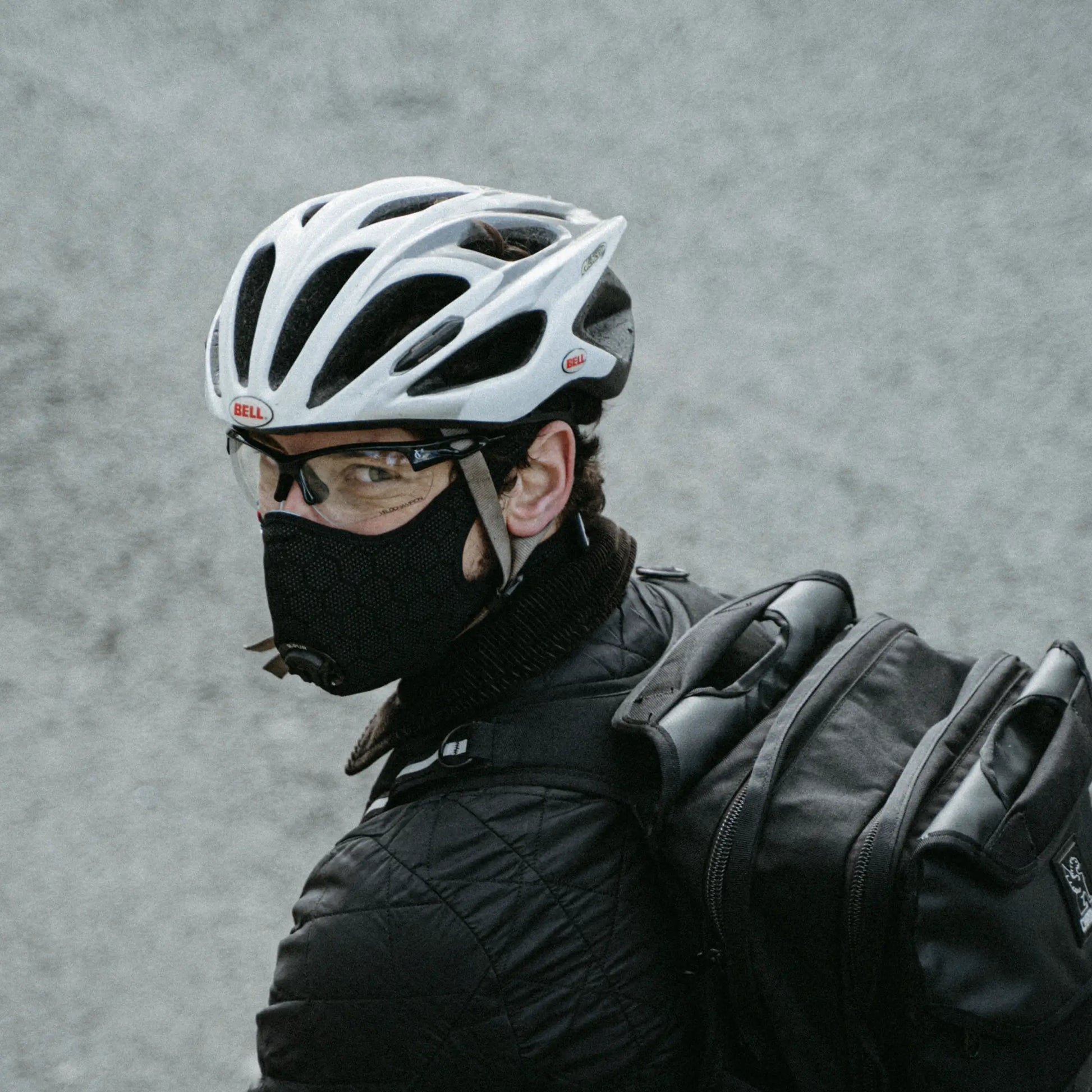 Cyclist on the road looking back, wearing a black R-PUR mask, white helmet, glasses and a black backpack.