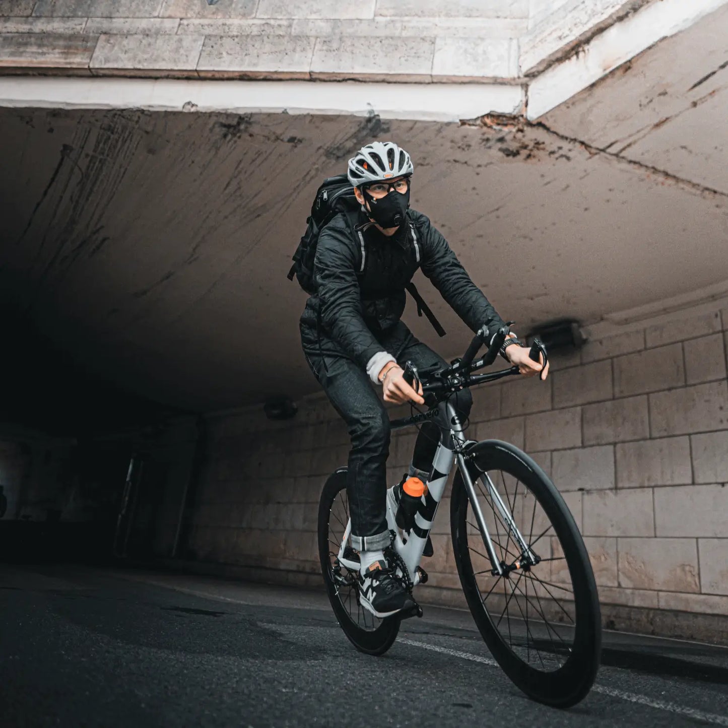 Cyclist wearing a black R-PUR cycling mask rides a road bike through an underpass with a backpack.